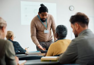 Black teacher talking to senior man during adult education training class in lecture hall.